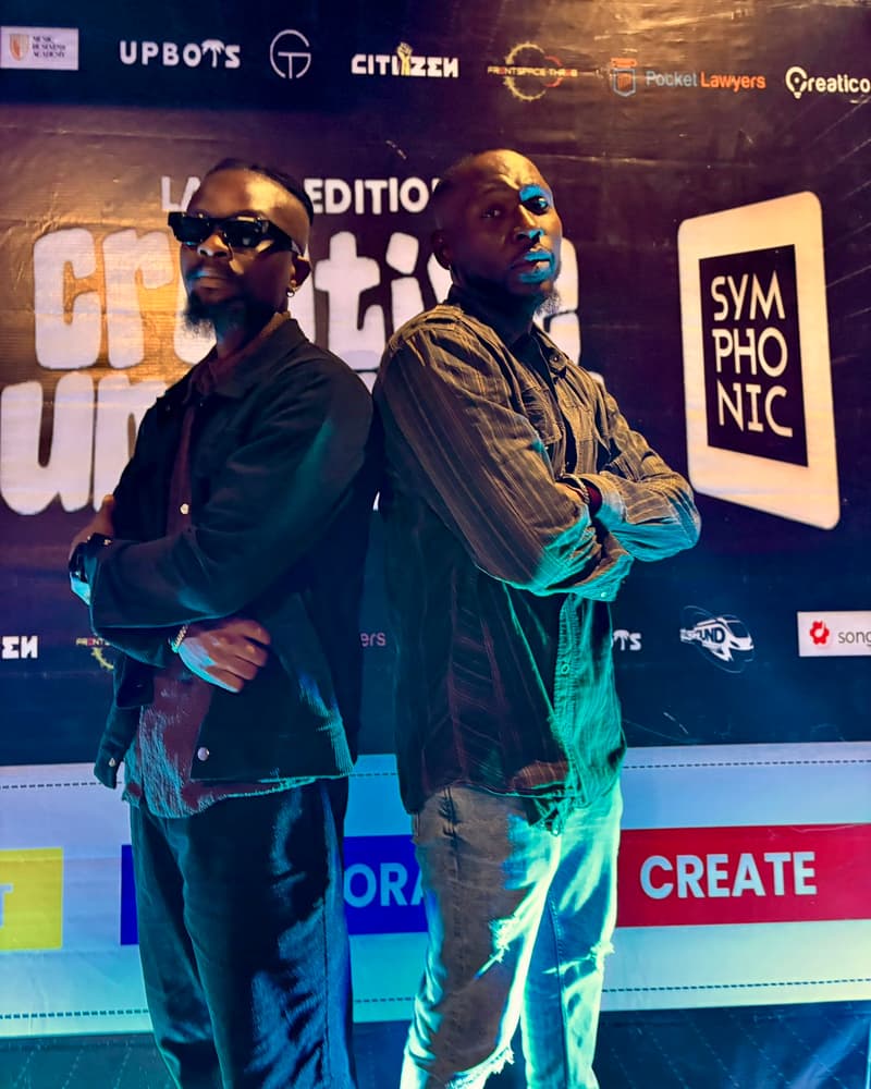 Two men standing back to back with their arms folded in front of the Creative Summit backdrop
